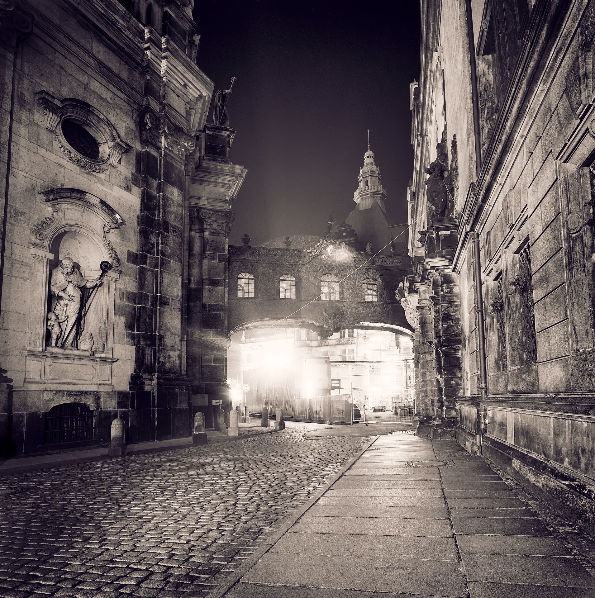 Cobbled baroque street at night between carved stone facades, lit archway and church tower in mist ahead