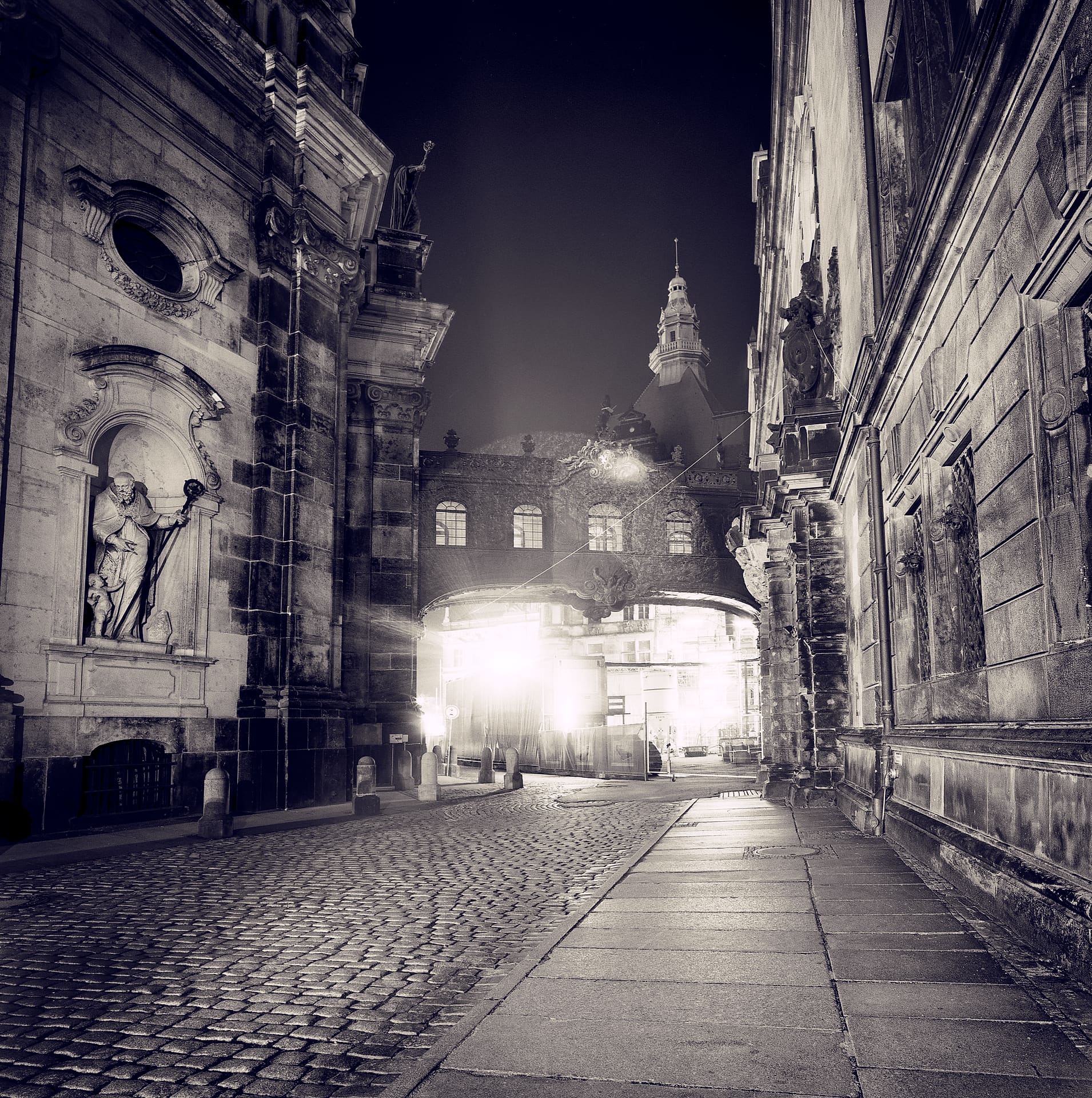 Cobbled baroque street at night between carved stone facades, lit archway and church tower in mist ahead