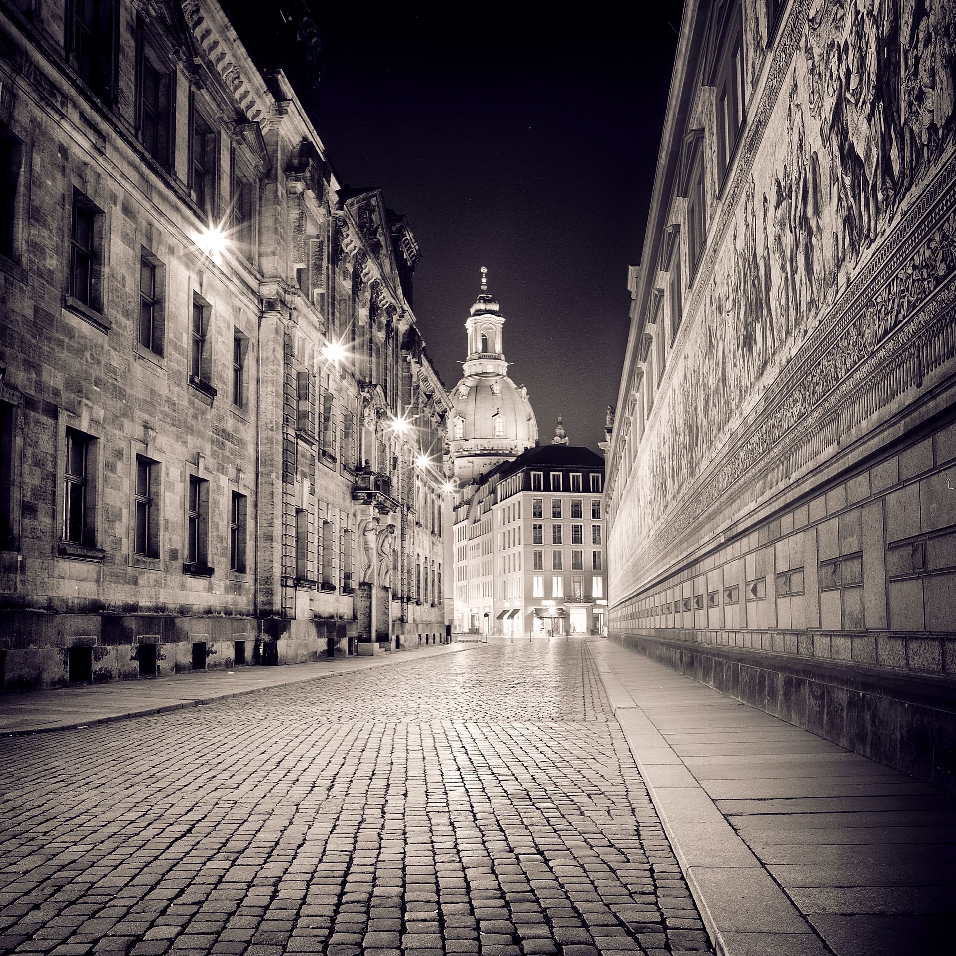 Empty cobbled street at night running toward the lit dome of the Frauenkirche, tiled Fürstenzug mural at right
