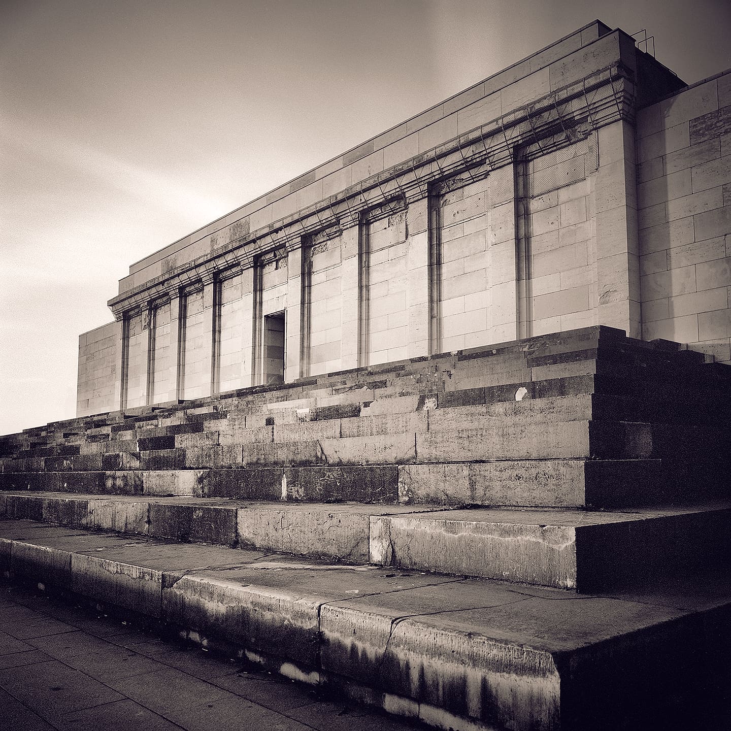 Weathered stone grandstand with pilastered colonnade seen along its broken tiered steps under flat grey sky