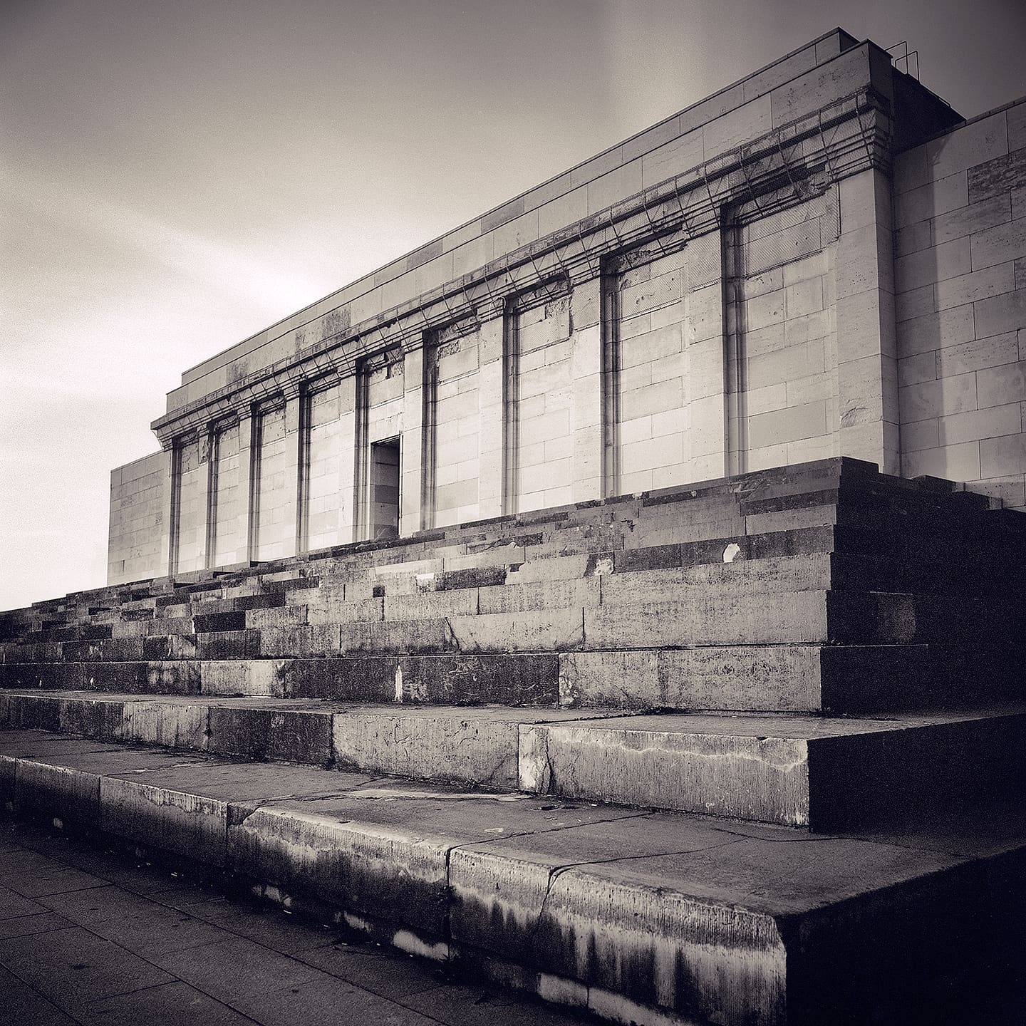 Weathered stone grandstand with pilastered colonnade seen along its broken tiered steps under flat grey sky
