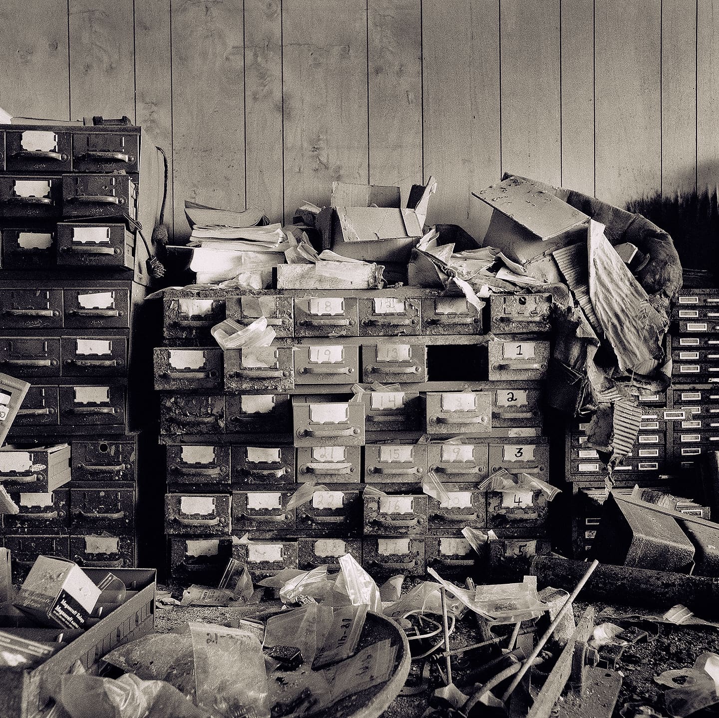 Stacked rusting card-index drawers against a wooden wall, papers, torn boxes and scattered rubbish across the floor