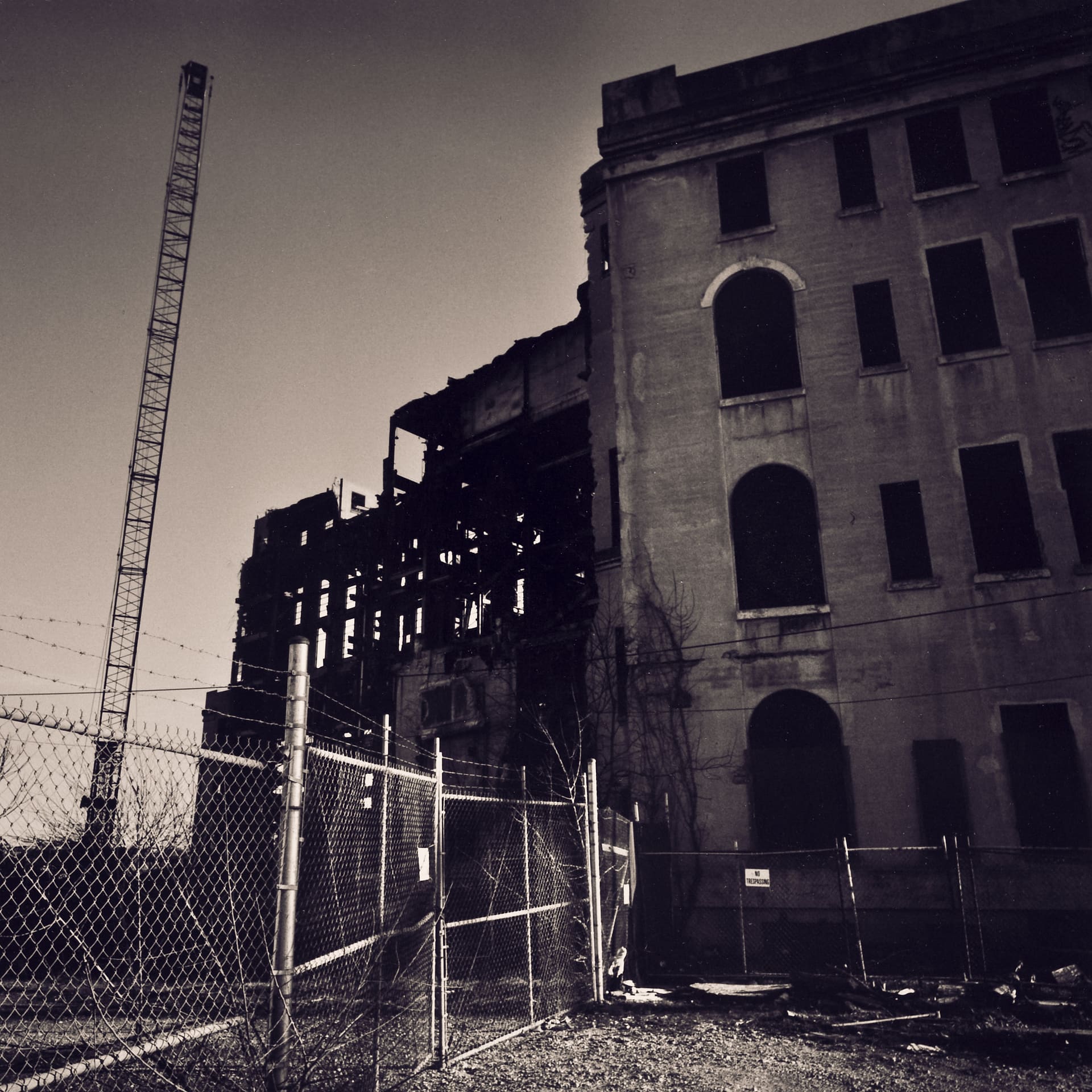 Gutted masonry power station behind chain-link and barbed-wire fencing, a lattice crane rising against a bare sky
