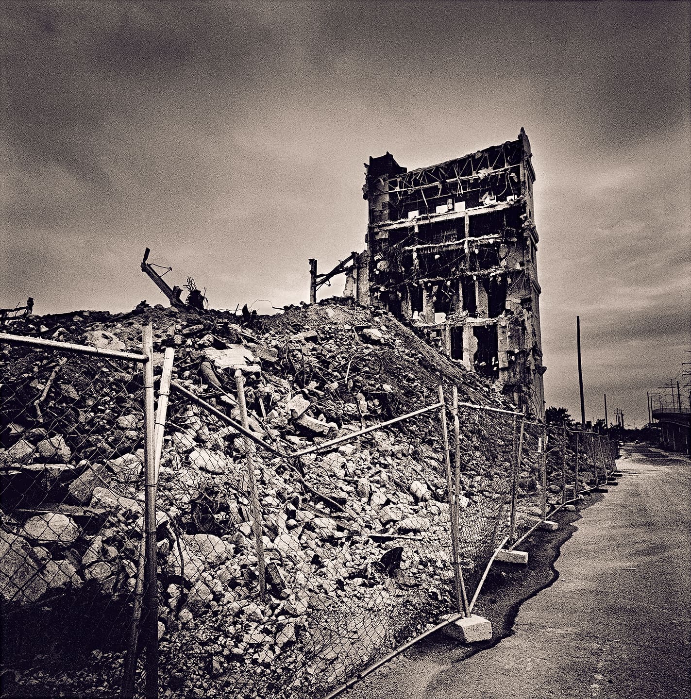 Half-demolished concrete tower stripped to its frame above a long rubble mound behind chain-link fencing beside an empty road