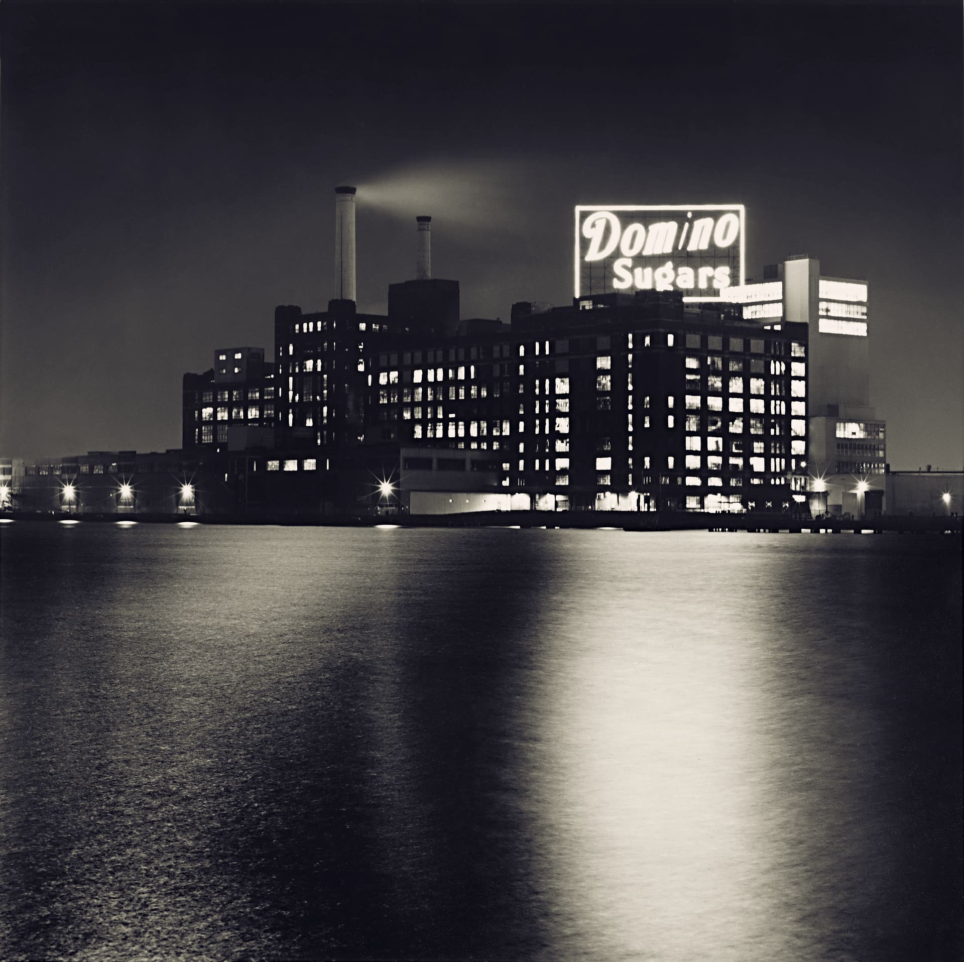 Illuminated Domino Sugars sign atop a lit waterfront refinery at night, its glow reflected in still harbour water