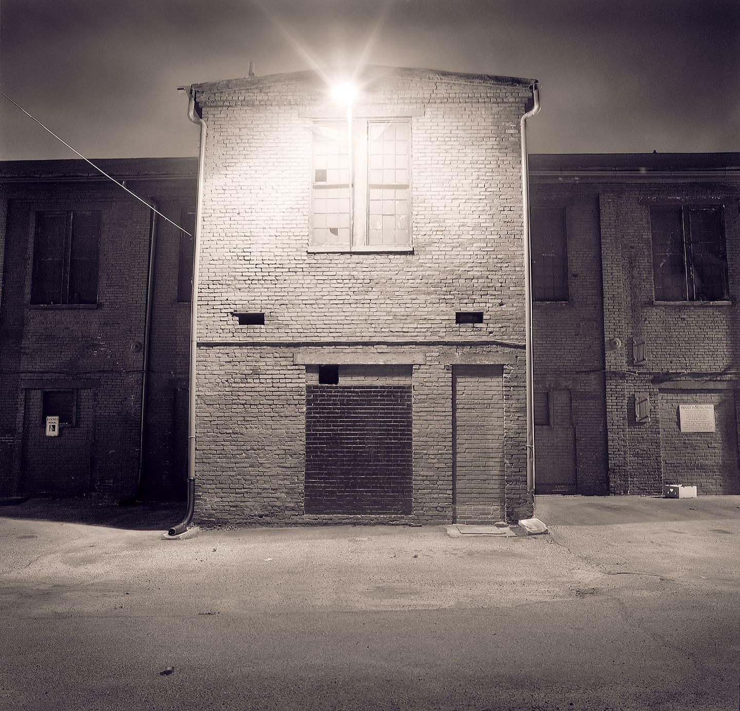 Small brick industrial building lit at night by a single flaring lamp above a broken upper window, bricked-up doorway below