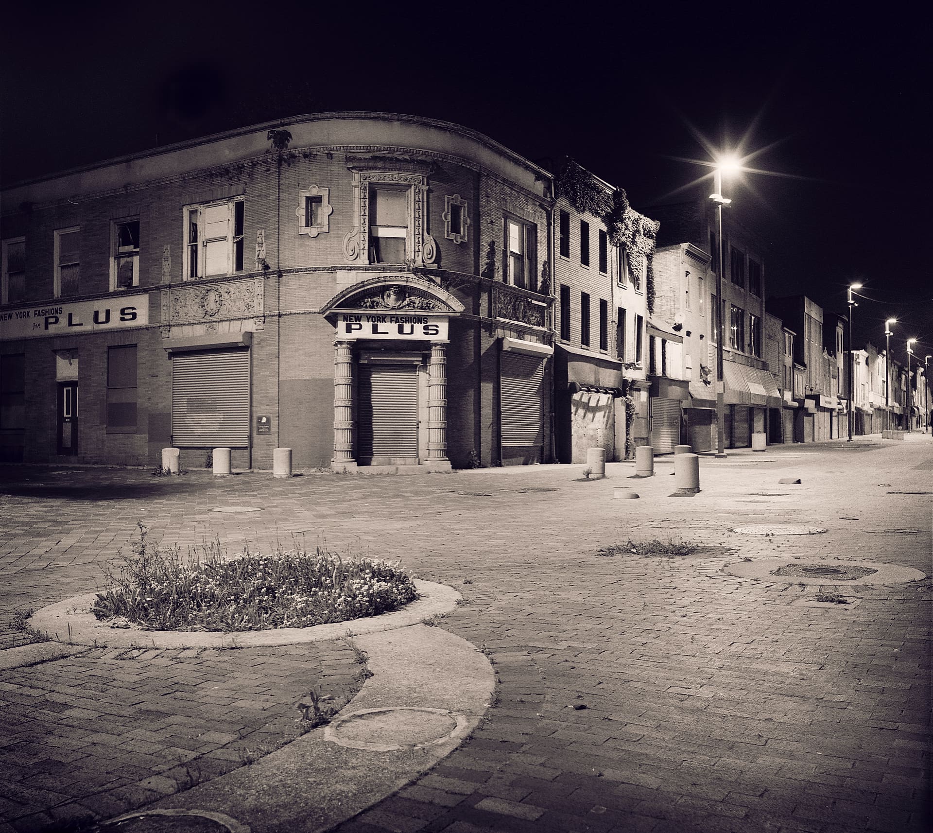 Empty brick pedestrian street at night, shuttered corner shopfront lettered New York Fashions Plus, weeds in a planter