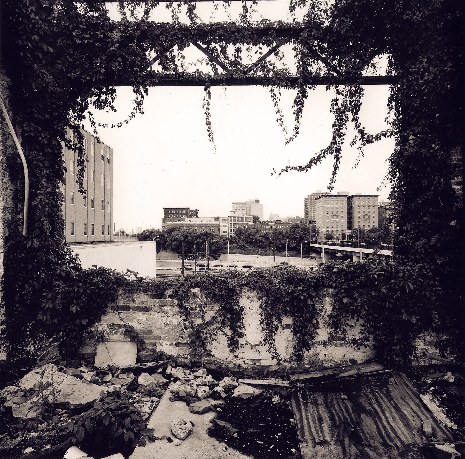 City skyline and elevated road framed through a ruined ivy-covered wall opening, rubble and rusted sheeting inside