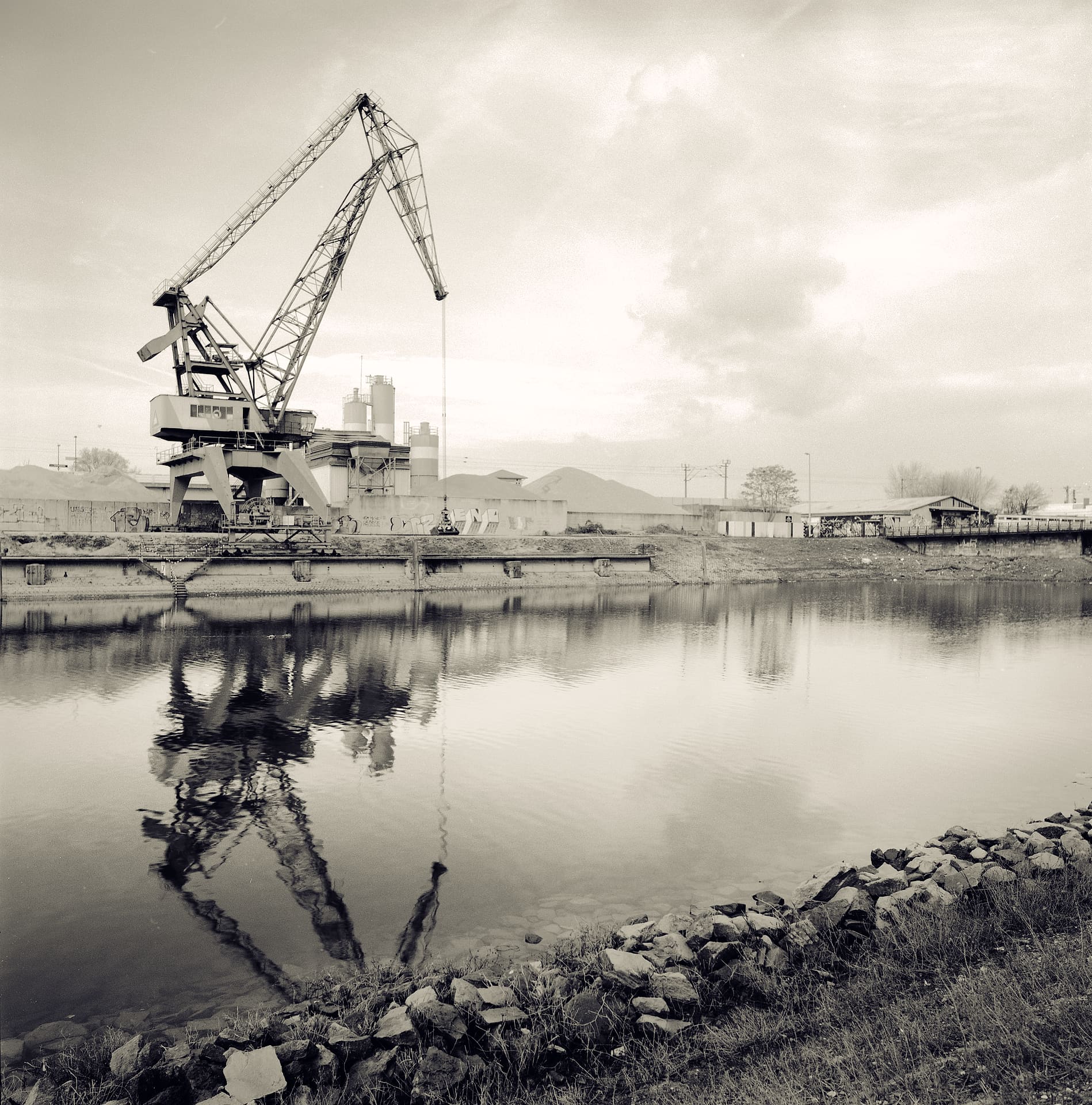 Disused harbour gantry crane and cement silos mirrored in still dock water, rip-rap stones along the near bank