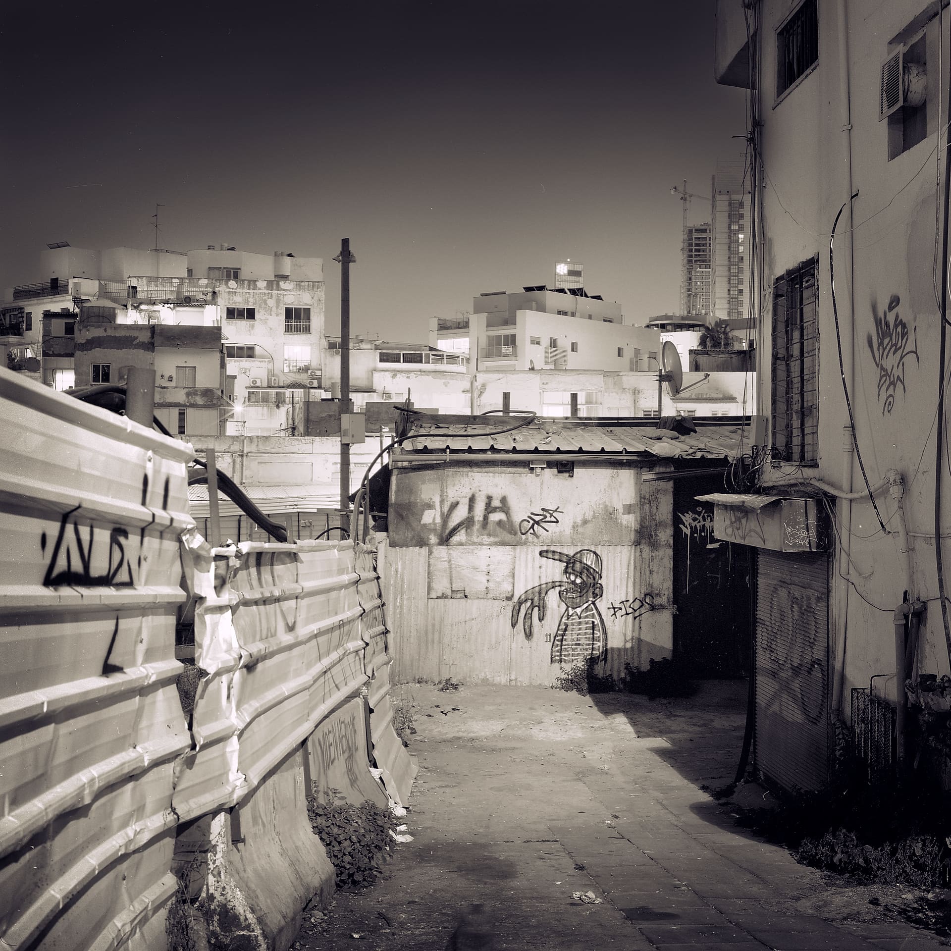 Dented corrugated hoarding beside a graffitied shed, apartment blocks and a construction crane rising behind at night