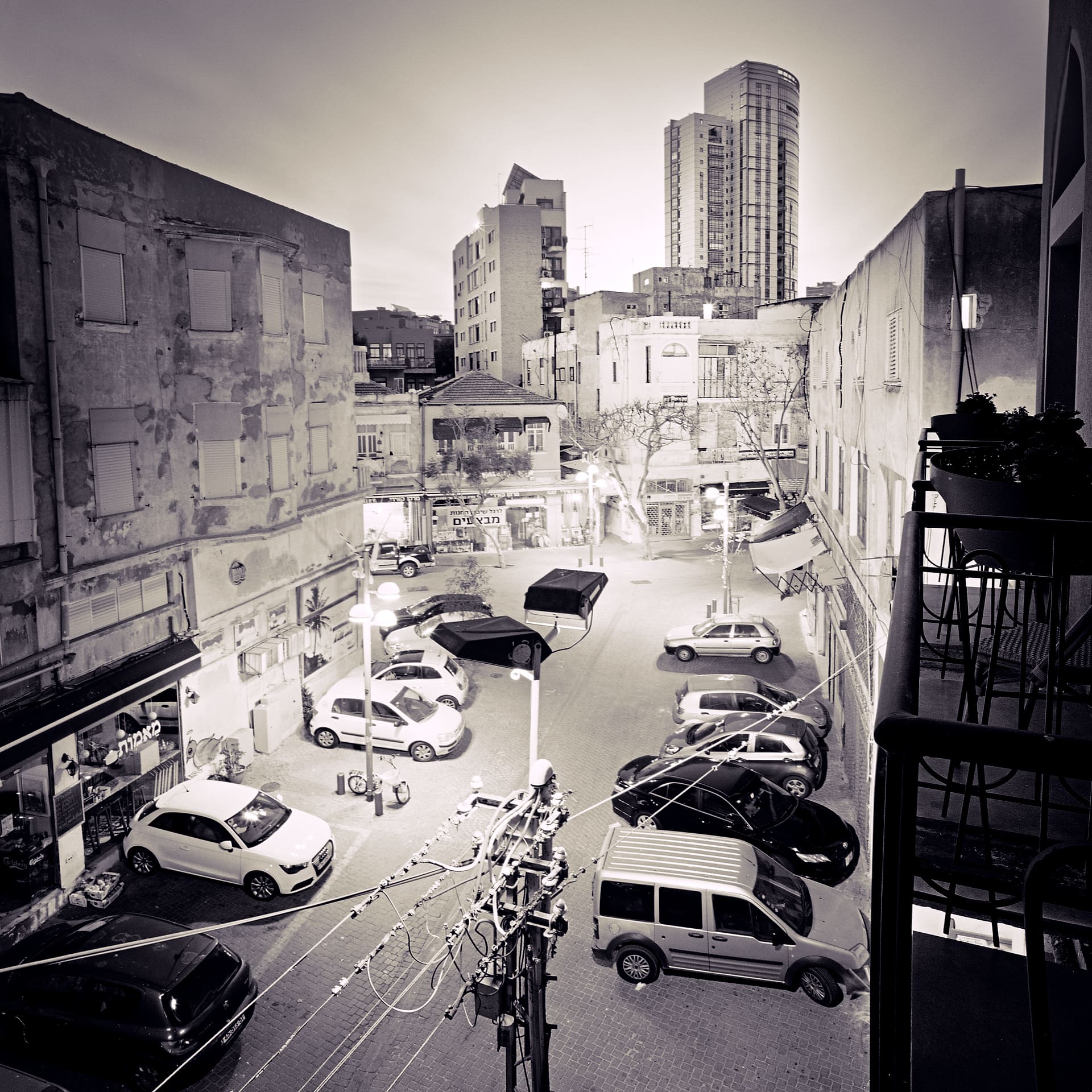 Balcony view down onto a parked car lot ringed by peeling apartment blocks, a round tower beyond, Hebrew shop signs