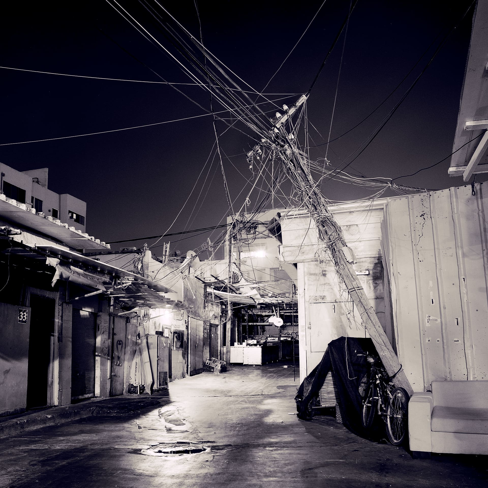 Tangle of overhead cables on a leaning utility pole above a wet market alley at night, bicycle and abandoned sofa