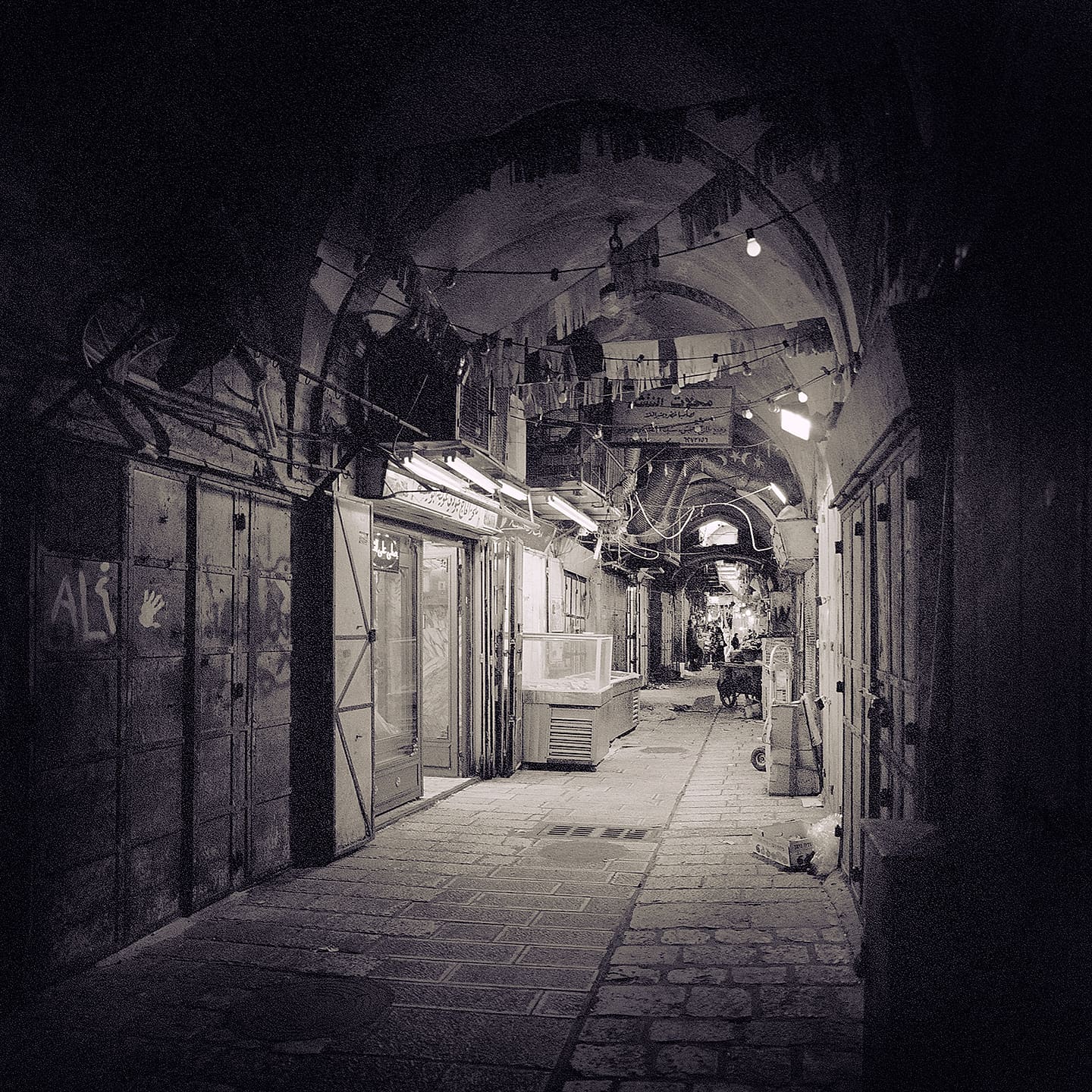 Vaulted stone bazaar lane at night, shuttered shopfronts, strung bulbs and Arabic signs, distant figures and a cat