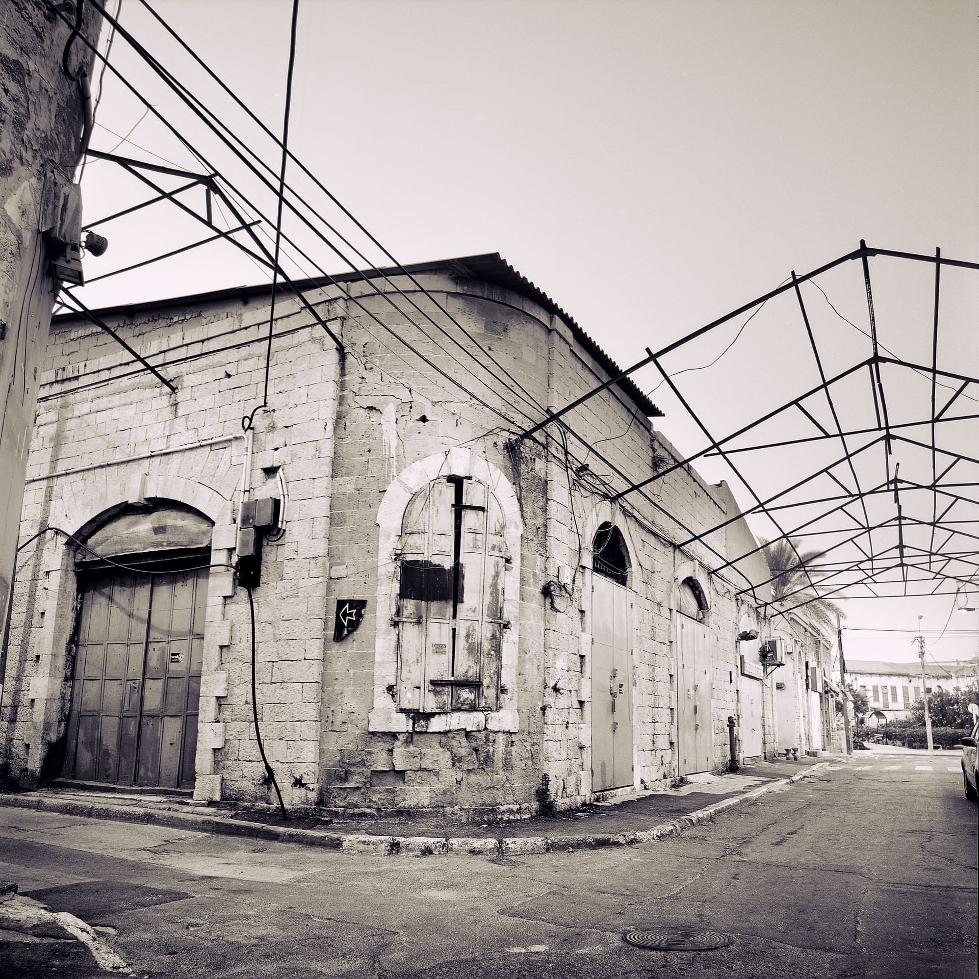 Corner of a weathered stone warehouse with shuttered arched doors, tangled overhead cables and a bare metal canopy frame
