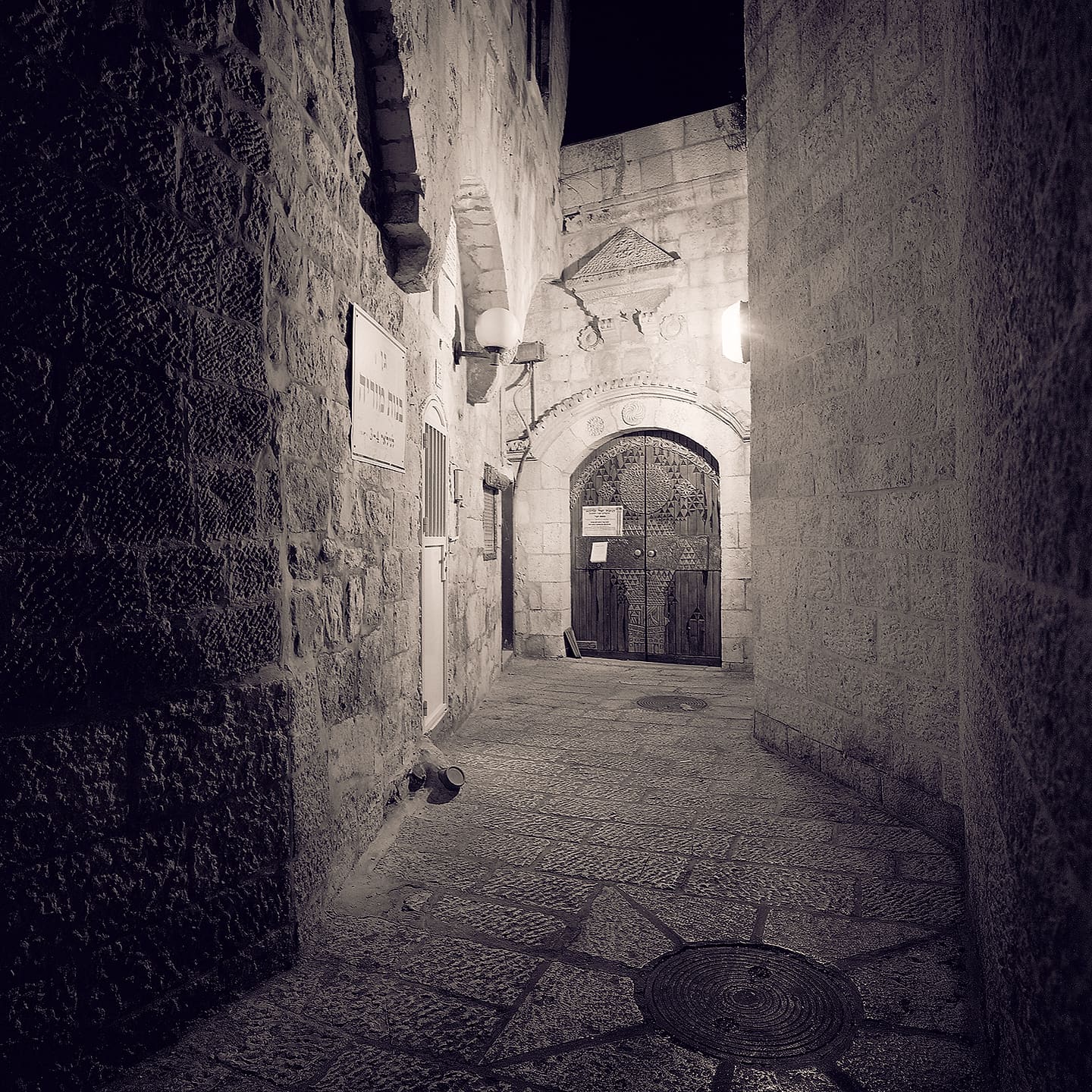 Narrow stone alley at night ending at a carved arched wooden door lit by wall lamps