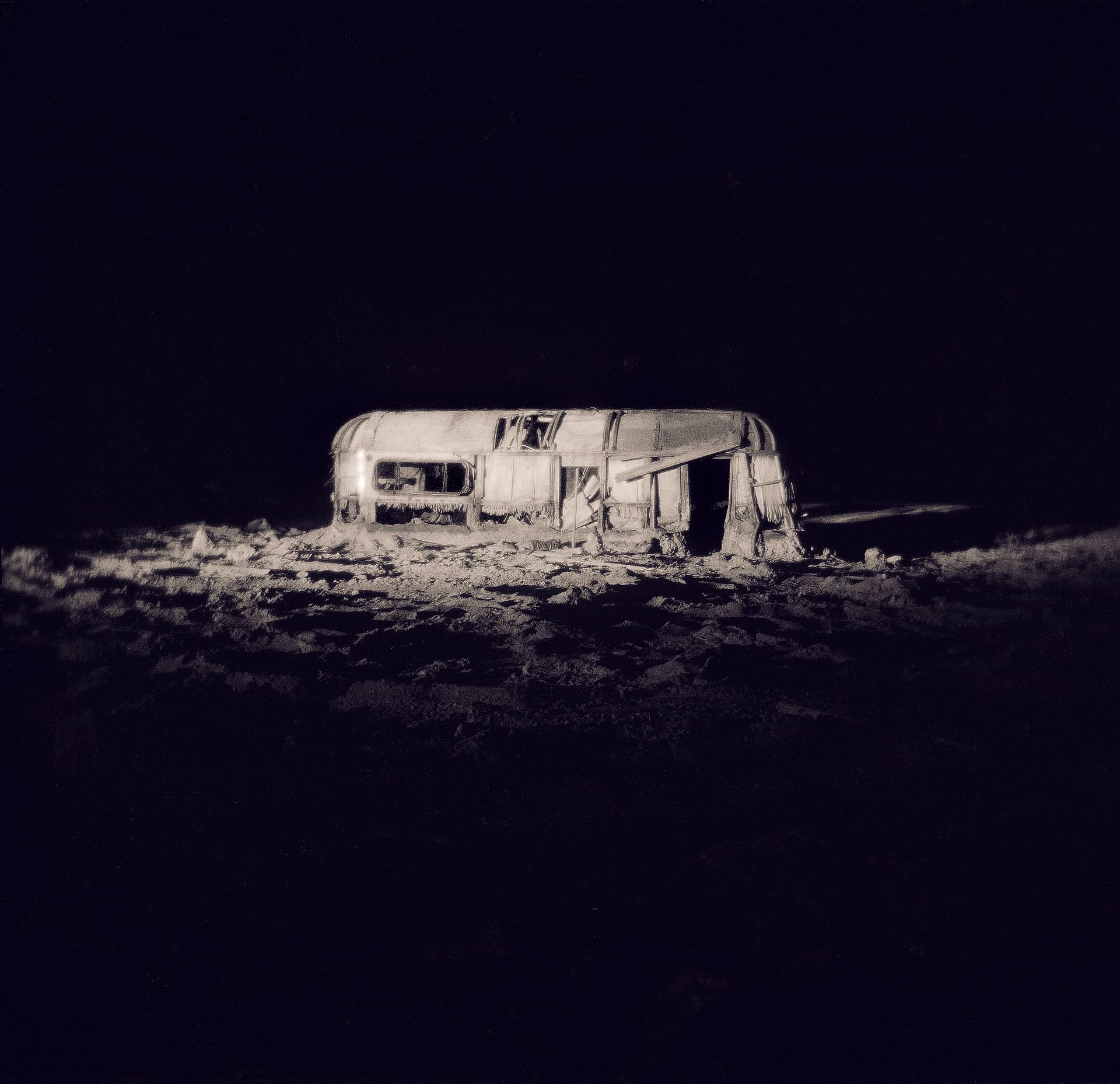 Wrecked caravan lit alone at night against total darkness, its shell and the ground around it glowing