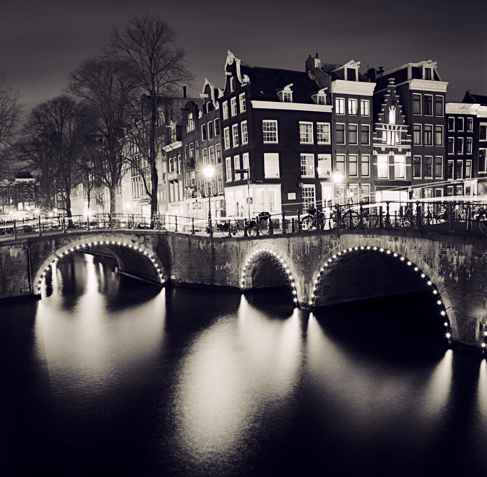 Lit canal bridge arches reflected in still water at night, gabled canal houses and moored bicycles behind