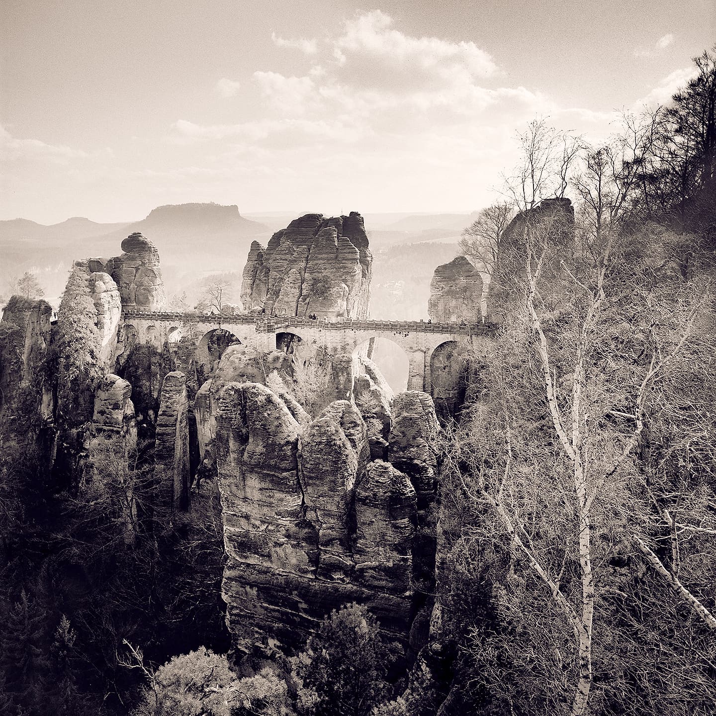 Stone arched bridge spanning between eroded sandstone pinnacles above a wooded valley, hazy hills beyond