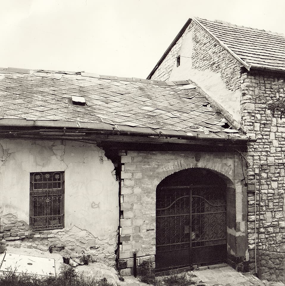 Crumbling stucco outbuilding with slate roof, ironwork gate and barred window beside a stone gable wall