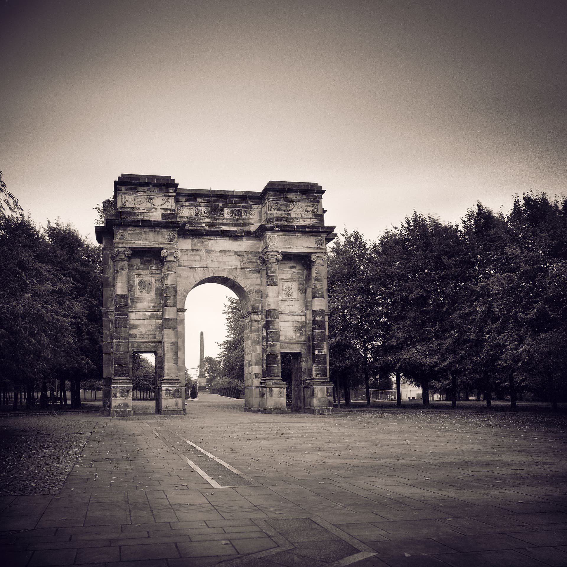 Weathered stone triumphal arch on an empty paved park avenue, flanked by trees, an obelisk beyond