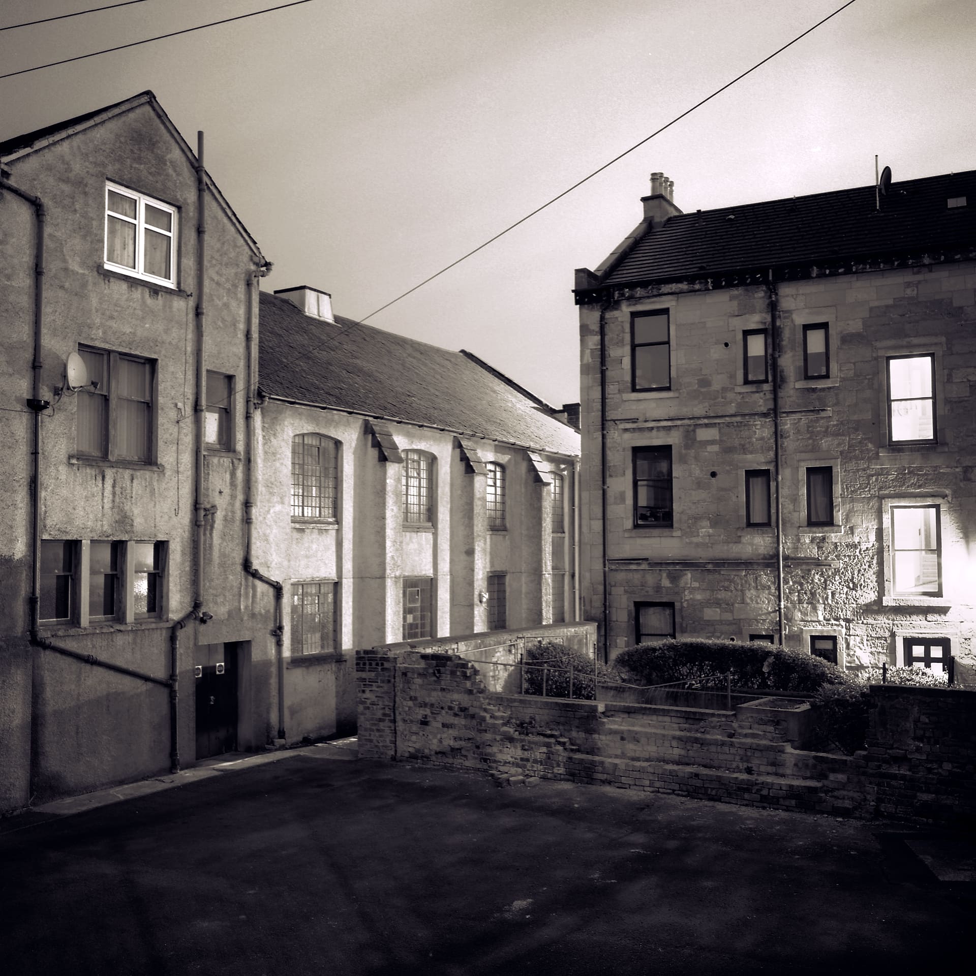Night view of stone tenement backs and a pitched-roof hall across an empty tarmac yard, overhead cables crossing the sky