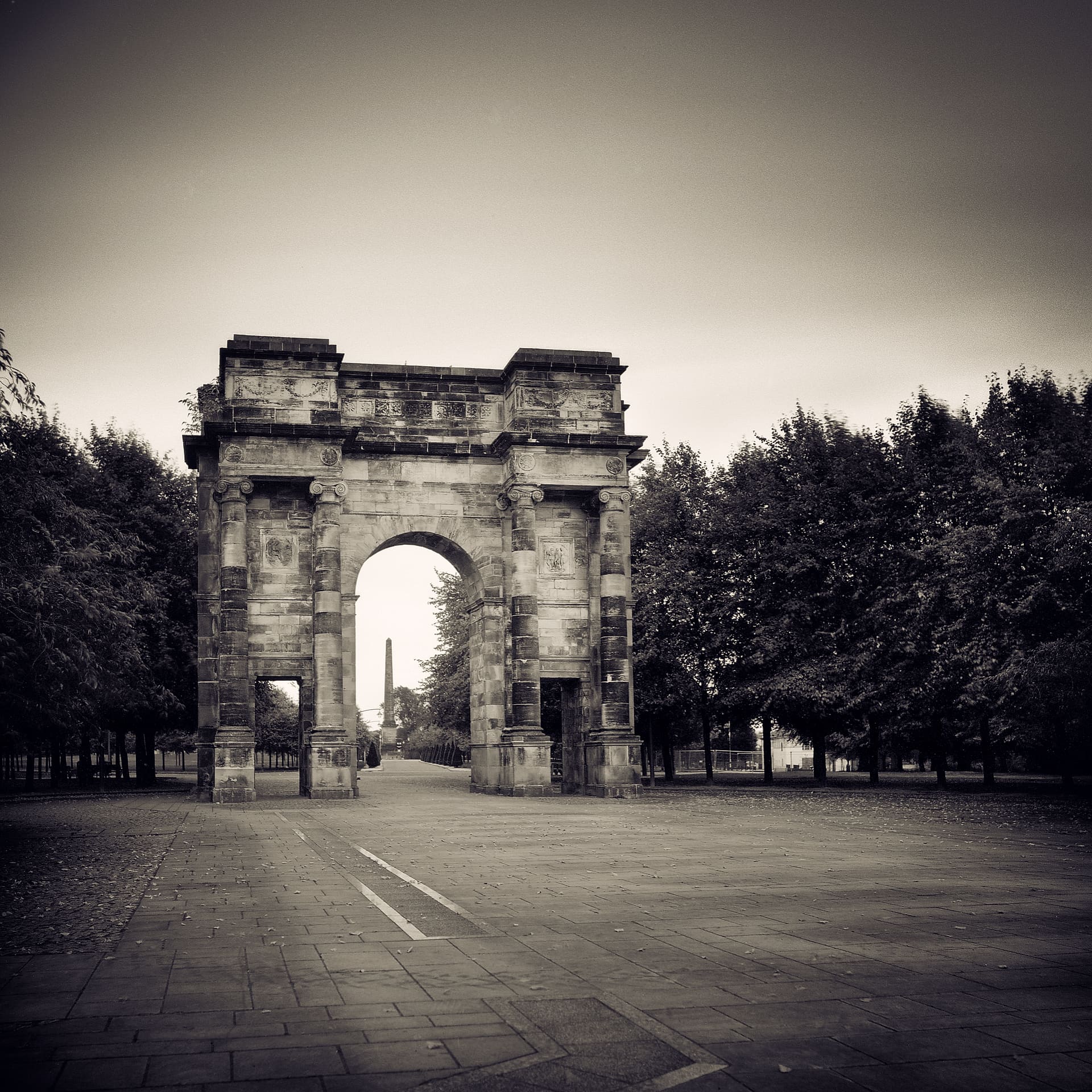 Weathered stone triumphal arch on an empty paved park avenue, flanked by trees, an obelisk beyond