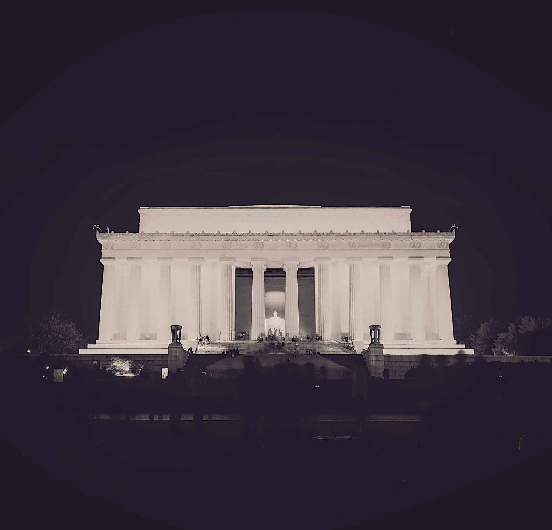 Floodlit marble colonnade of the Lincoln Memorial seen head-on at night, the seated statue lit within, blurred crowd on the steps and darkness above