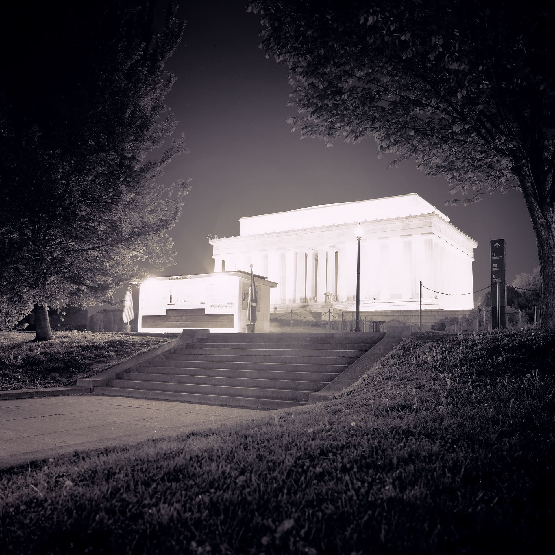 Floodlit colonnaded marble memorial glowing at night behind stone steps, dark trees framing both sides