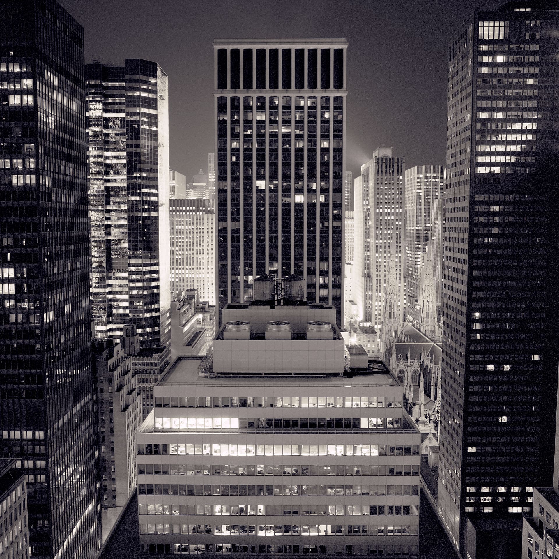Midtown office towers at night seen from above, lit windows and rooftop mechanical plant, cathedral spires at right