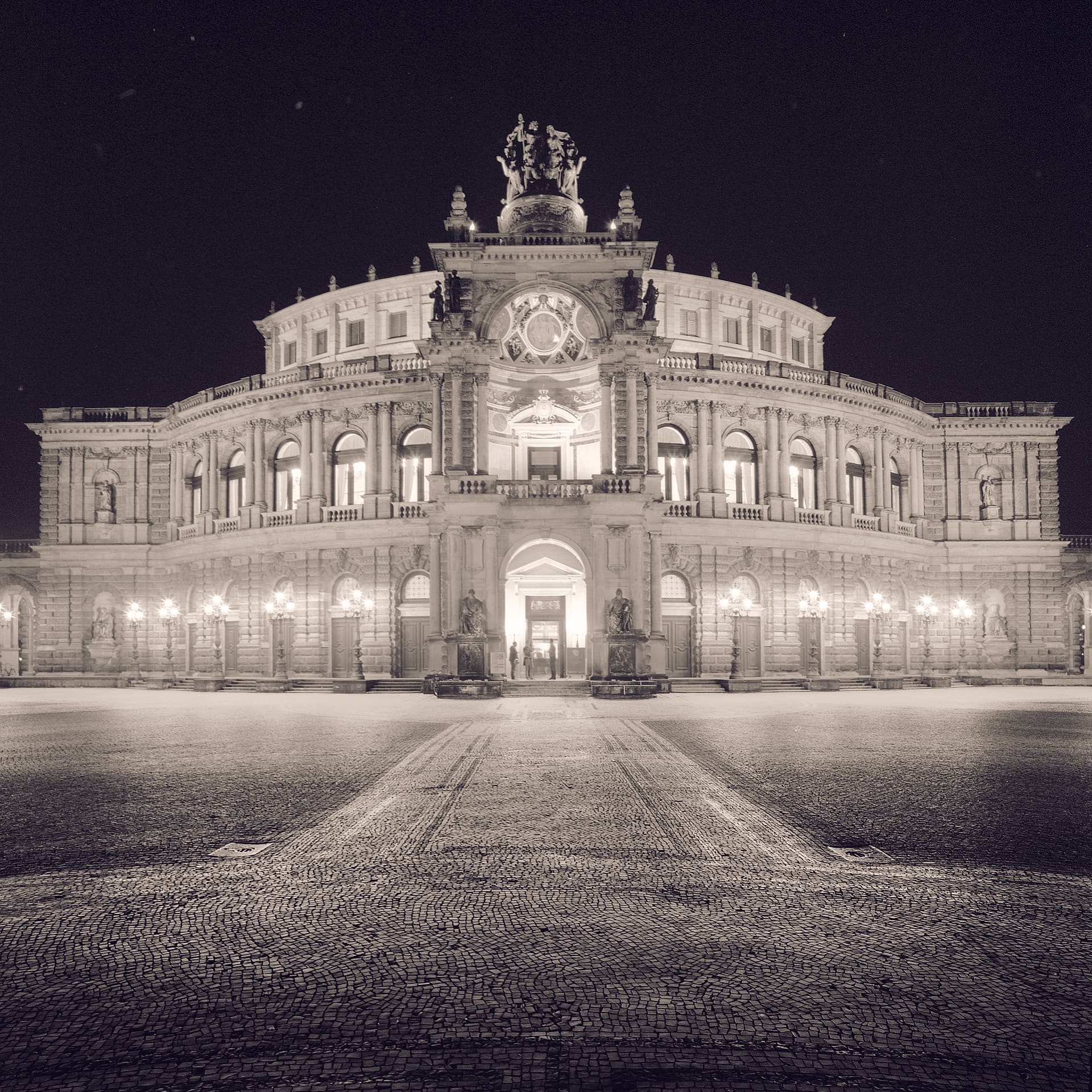 Floodlit neoclassical opera house with quadriga sculpture above its entrance, empty cobbled square in the foreground at night