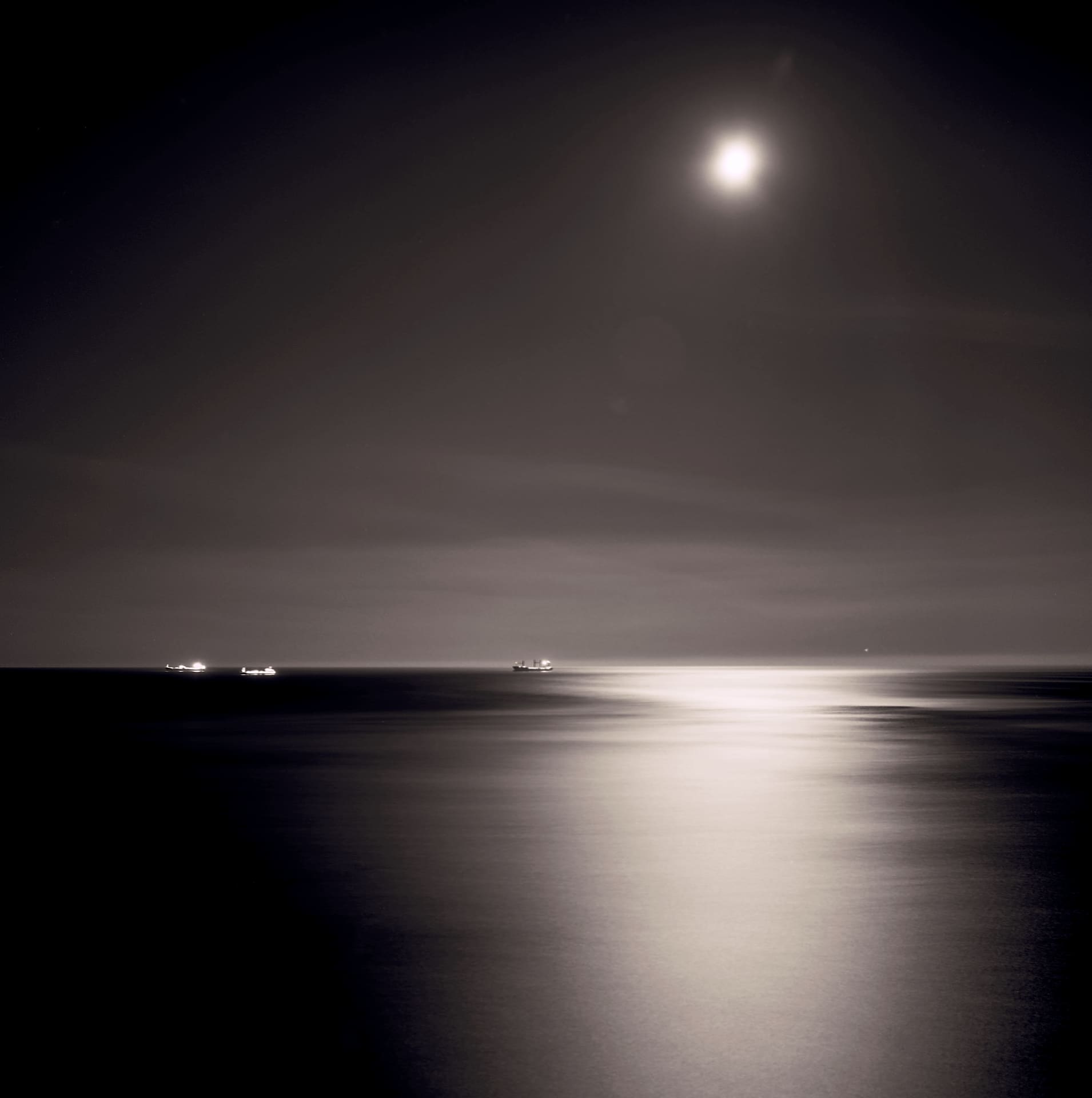 Full moon over open sea at night, its light laid in a broad streak across smoothed water, three lit ships on the horizon