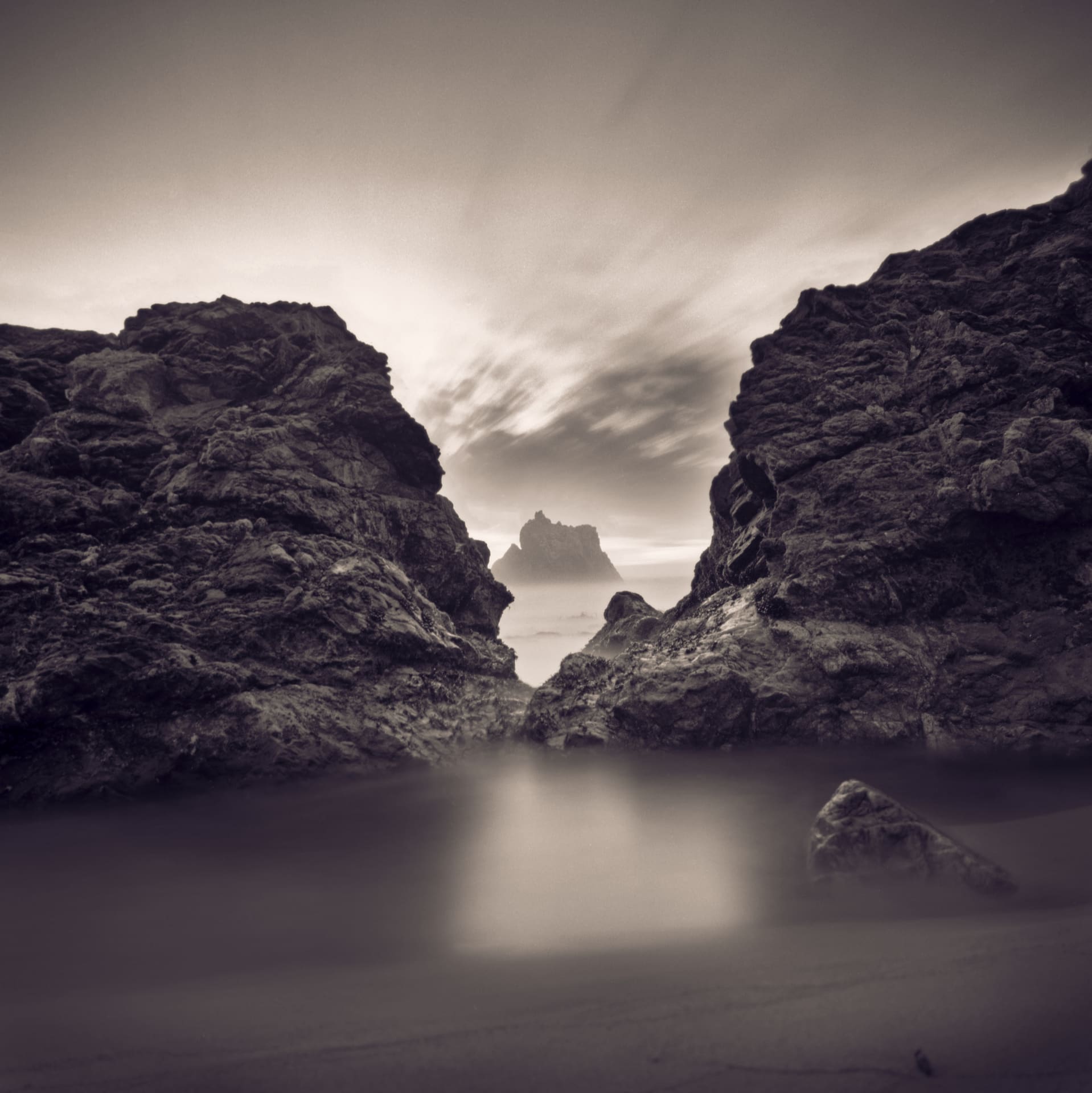 Sea stack framed between dark headland rocks, long exposure smoothing surf and streaking cloud overhead