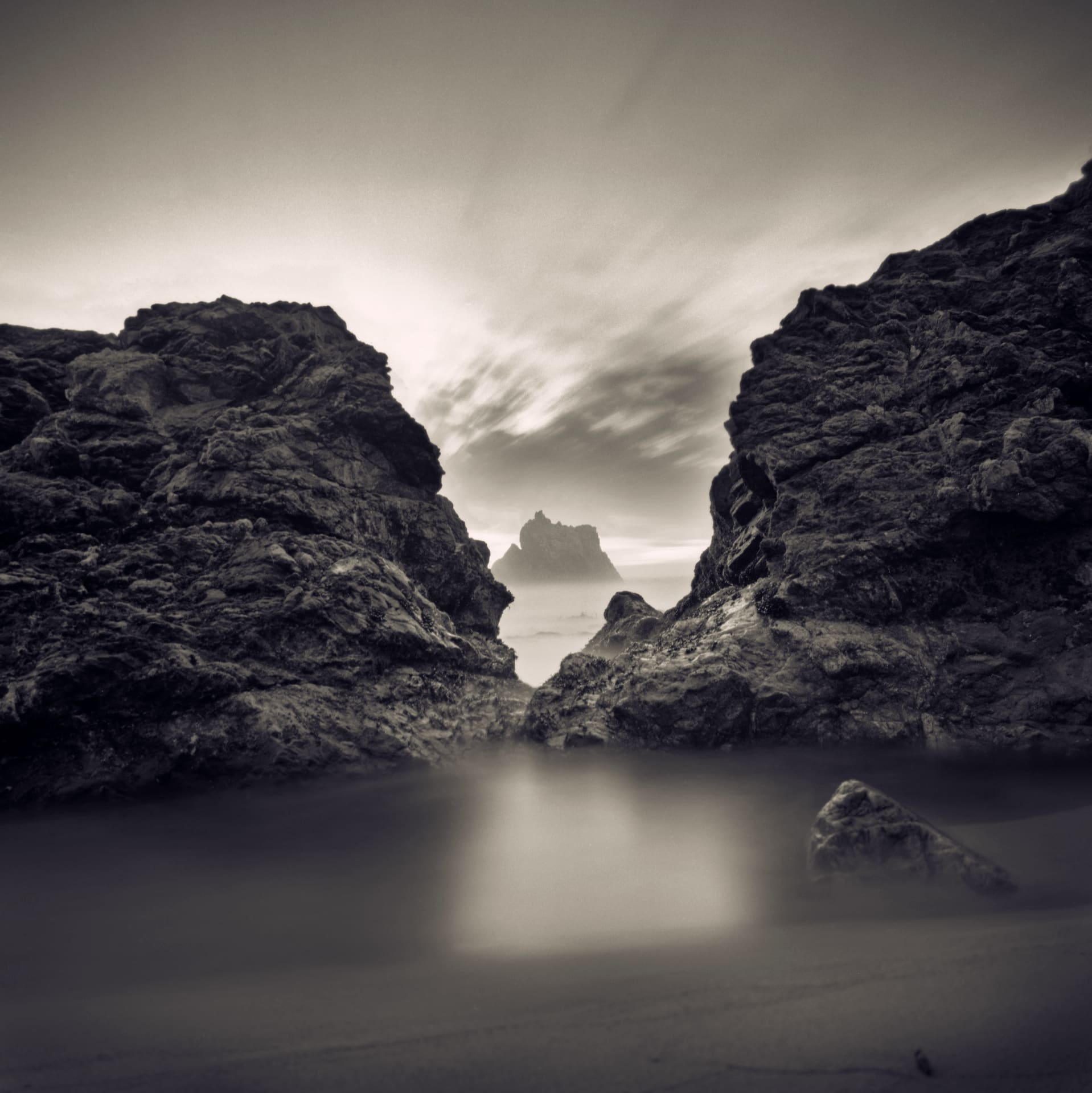 Sea stack framed between dark headland rocks, long exposure smoothing surf and streaking cloud overhead
