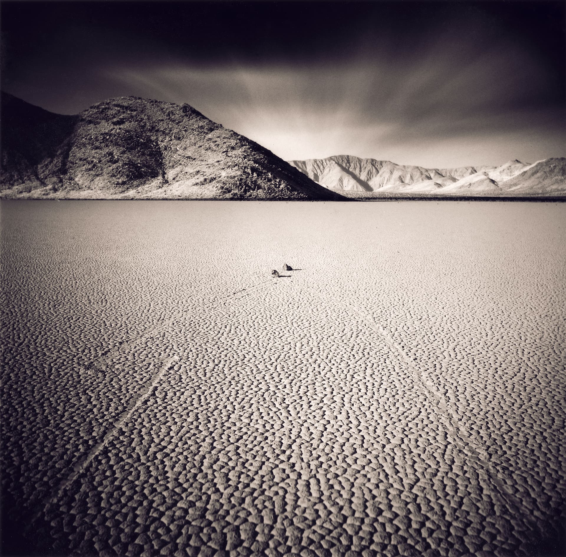 Cracked dry lakebed with a sailing stone and its long track, dark mountains and streaked cloud above