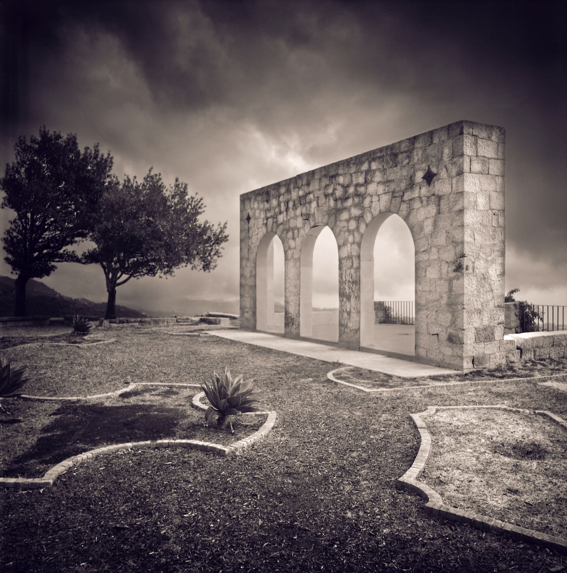 Freestanding stone wall pierced by three arches on a clipped hillside garden, storm cloud massing behind